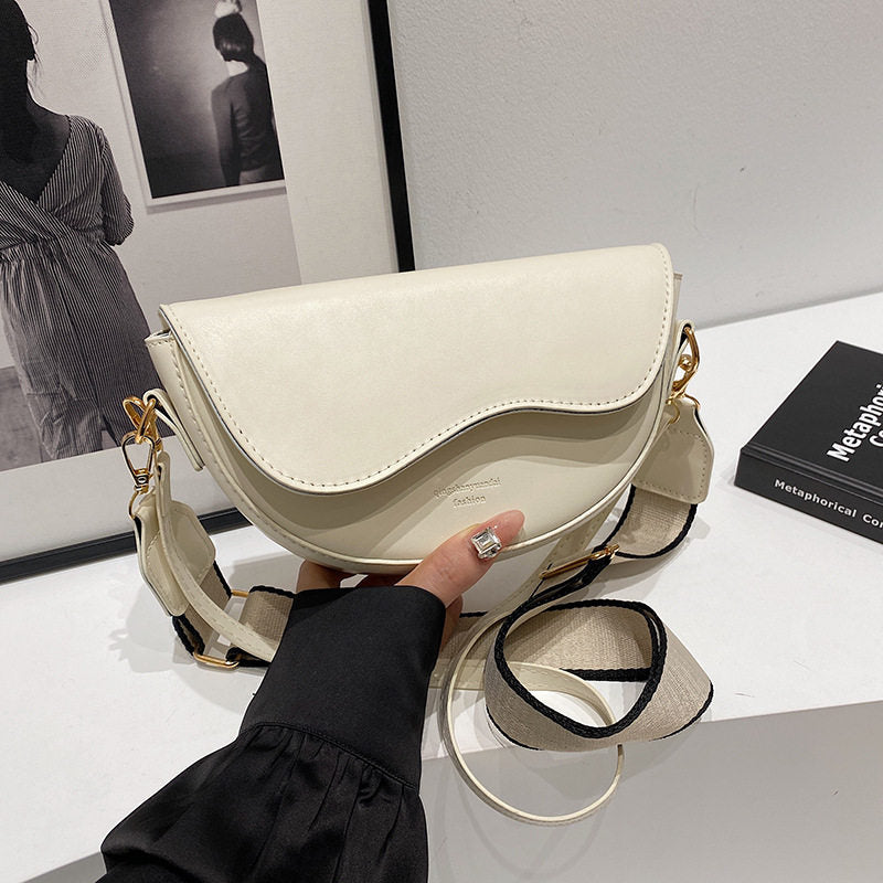 Beige handbag held by a person on a white surface with a black book and framed picture in the background.